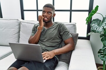 Young african american man using laptop at home sitting on the sofa looking stressed and nervous with hands on mouth biting nails. anxiety problem.