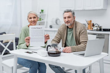 Pensioners holding contract and looking at camera near devices and coffee in kitchen.
