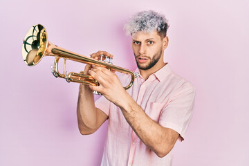 Young hispanic man with modern dyed hair playing trumpet clueless and confused expression. doubt...