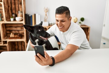 Young hispanic man make selfie by the smartphone sitting on the table with dog at home.