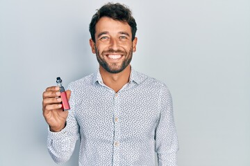 Handsome man with beard football reporter holding electronic cigarette looking positive and happy standing and smiling with a confident smile showing teeth