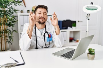 Young doctor working at the clinic using computer laptop gesturing finger crossed smiling with hope and eyes closed. luck and superstitious concept.