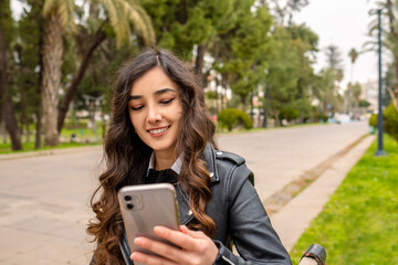 Pretty young woman talking with friends on phone video call. Checking social media holding smartphone at outdoor. Smiling young woman using mobile phone app playing game, shopping online on a park