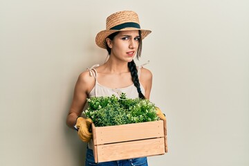 Young hispanic woman holding green plant pot clueless and confused expression. doubt concept.