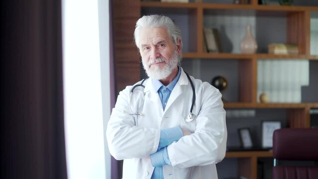 Portrait elder gray-haired experienced senior doctor standing in a modern hospital clinic in his office, looking at camera. crossed arms. confident mature older physician general practitioner smiling