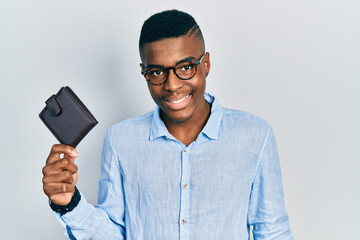 Young african american man wearing glasses holding leather wallet looking positive and happy standing and smiling with a confident smile showing teeth