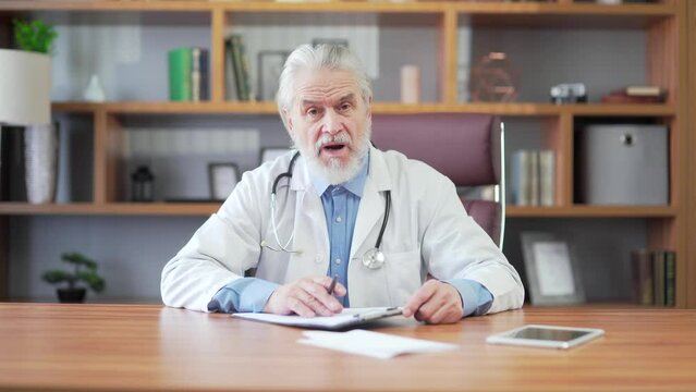 Portrait senior experienced male doctor consulting on video call looking at camera in a hospital clinic. Mature gray haired older woman gives advice to the patient Webcam view POV Medical Consultation