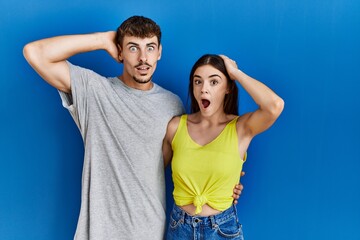 Young hispanic couple standing together over blue background crazy and scared with hands on head, afraid and surprised of shock with open mouth