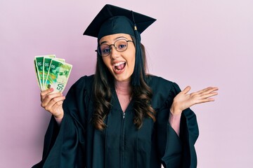 Young hispanic woman wearing graduation uniform holding israel shekels banknotes celebrating achievement with happy smile and winner expression with raised hand