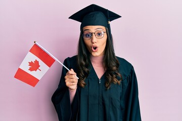 Young hispanic woman wearing graduation uniform holding canada flag scared and amazed with open mouth for surprise, disbelief face