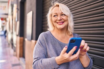 Middle age blonde woman smiling confident using smartphone at street