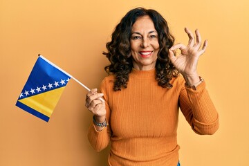 Middle age hispanic woman holding bosnia herzegovina flag doing ok sign with fingers, smiling friendly gesturing excellent symbol