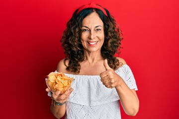 Middle age hispanic woman holding potato chip smiling happy and positive, thumb up doing excellent and approval sign