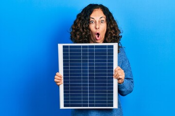 Middle age hispanic woman holding photovoltaic solar panel afraid and shocked with surprise and...
