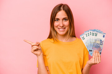 Young caucasian woman holding banknotes isolated on pink background smiling and pointing aside,...