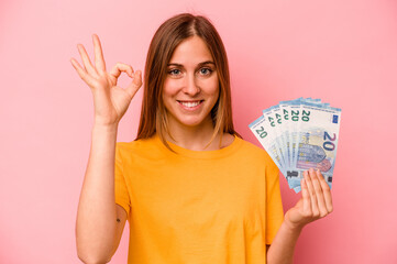 Young caucasian woman holding banknotes isolated on pink background cheerful and confident showing ok gesture.