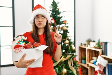 Young brunette woman standing by christmas tree holding decoration pointing up looking sad and upset, indicating direction with fingers, unhappy and depressed.