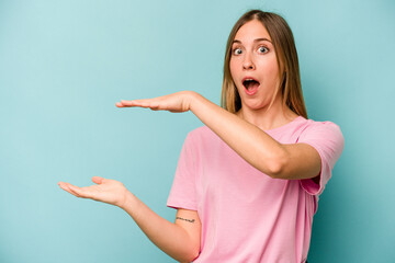 Young caucasian woman isolated on blue background shocked and amazed holding a copy space between hands.