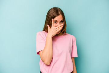 Young caucasian woman isolated on blue background blink at the camera through fingers, embarrassed covering face.