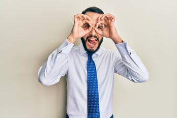 Young man with beard wearing business tie doing ok gesture like binoculars sticking tongue out, eyes looking through fingers. crazy expression.