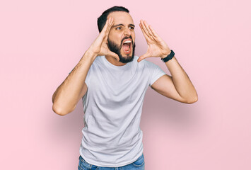 Young man with beard wearing casual white t shirt shouting angry out loud with hands over mouth