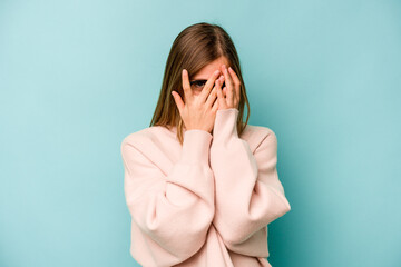 Young caucasian woman isolated on blue background blink at the camera through fingers, embarrassed covering face.