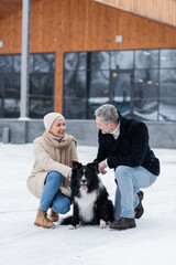Happy senior woman in winter outfit looking at husband near border collie on snow outdoors.
