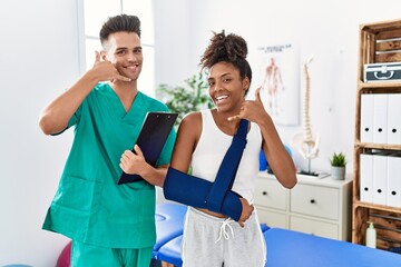 Physiotherapist working with patient wearing arm on sling at rehabilitation clinic smiling doing phone gesture with hand and fingers like talking on the telephone. communicating concepts.