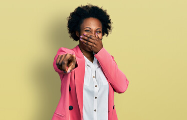 African american woman with afro hair wearing business jacket laughing at you, pointing finger to the camera with hand over mouth, shame expression