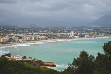 Open view of the landscape and the coast of Alfaz del Pi, with pine trees in the foreground and the Mediterranean Sea, the beach, the buildings of L'Albir and the mountains in the background.