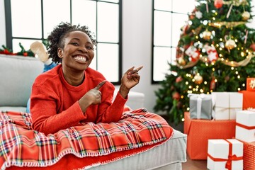 Young african american woman lying on the sofa by christmas tree smiling and looking at the camera pointing with two hands and fingers to the side.