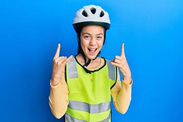 Beautiful brunette little girl wearing bike helmet and reflective vest shouting with crazy expression doing rock symbol with hands up. music star. heavy concept.