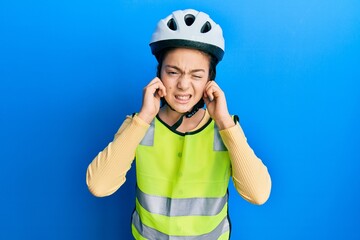 Beautiful brunette little girl wearing bike helmet and reflective vest covering ears with fingers with annoyed expression for the noise of loud music. deaf concept.