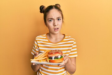 Beautiful brunette little girl eating a tasty classic burger with fries clueless and confused...