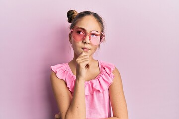 Beautiful brunette little girl wearing heart shaped sunglasses thinking concentrated about doubt...