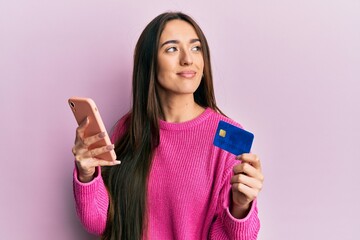 Young hispanic girl holding smartphone and credit card smiling looking to the side and staring away...