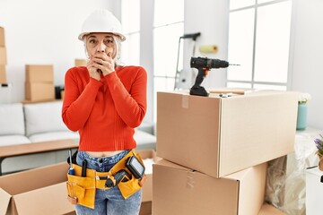 Middle age grey-haired woman wearing hardhat standing at new home shocked covering mouth with hands for mistake. secret concept.