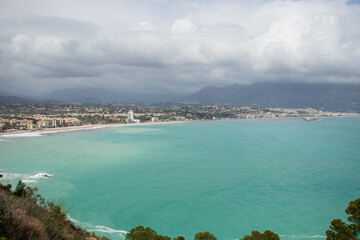 View of the small bay formed by the Racó Albir beach and Cap Blanc beach, in Alicante, during a cloudy day. Turquoise water of the Mediterranean Sea and mountains in the background.