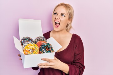 Young blonde woman holding tasty colorful doughnuts angry and mad screaming frustrated and furious, shouting with anger looking up.