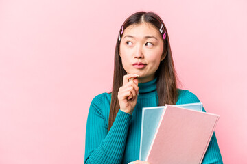 Obraz premium Young asian student woman holding books isolated on pink background looking sideways with doubtful and skeptical expression.