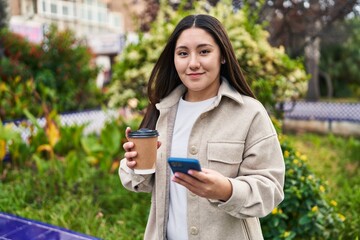 Young hispanic woman using smartphone drinking coffee at park