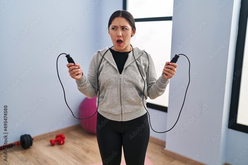 Poster young latin woman jumping with skipping rope angry and mad screaming frustrated and furious, shoutin