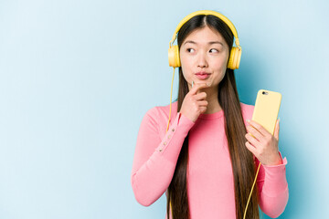 Young asian woman listening to music isolated on blue background looking sideways with doubtful and skeptical expression.