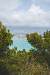 Fototapeta na wymiar Landscape of the coast of Alicante between pine trees of an intense green color on a cloudy day. Surroundings of the Costa Blanca, with the sea and the town of Albir and Alfaz del Pi on the horizon.