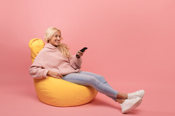 Television programming. Happy lady with remote control sitting in beanbag chair, switching channels, pink background