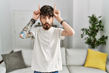Hispanic man with beard at the living room at home doing funny gesture with finger over head as bull horns