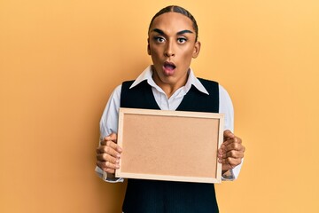 Hispanic man wearing make up and long hair holding empty corkboard afraid and shocked with surprise and amazed expression, fear and excited face.