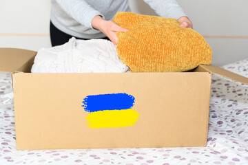 Woman puts winter clothes in a cardboard box with the colors of the Ukrainian flag, yellow and blue, drawn on it. Concept of donation to Ukrainian refugees