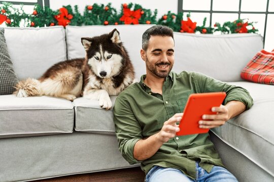 Young hispanic man using touchpad sitting on sofa with dog by christmas decor at home - Powered by Adobe