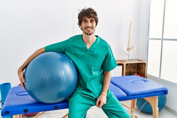Young hispanic physiotherapist man holding pilates ball at pain recovery clinic looking positive and happy standing and smiling with a confident smile showing teeth